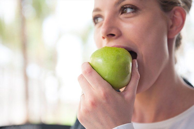 Person biting a green apple while looking away in a bright indoor setting