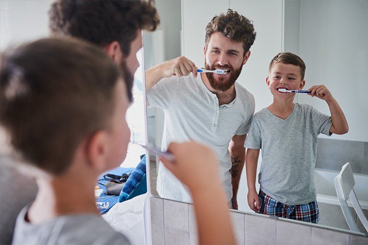 Man brushing teeth at bathroom mirror, smiling child beside him, both in gray shirts