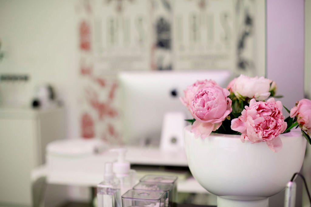 Eine weiße Schale mit rosa Blumen steht auf einem Schminktisch; im Hintergrund sind ein verschwommener Spiegel und Flaschen zu sehen.
