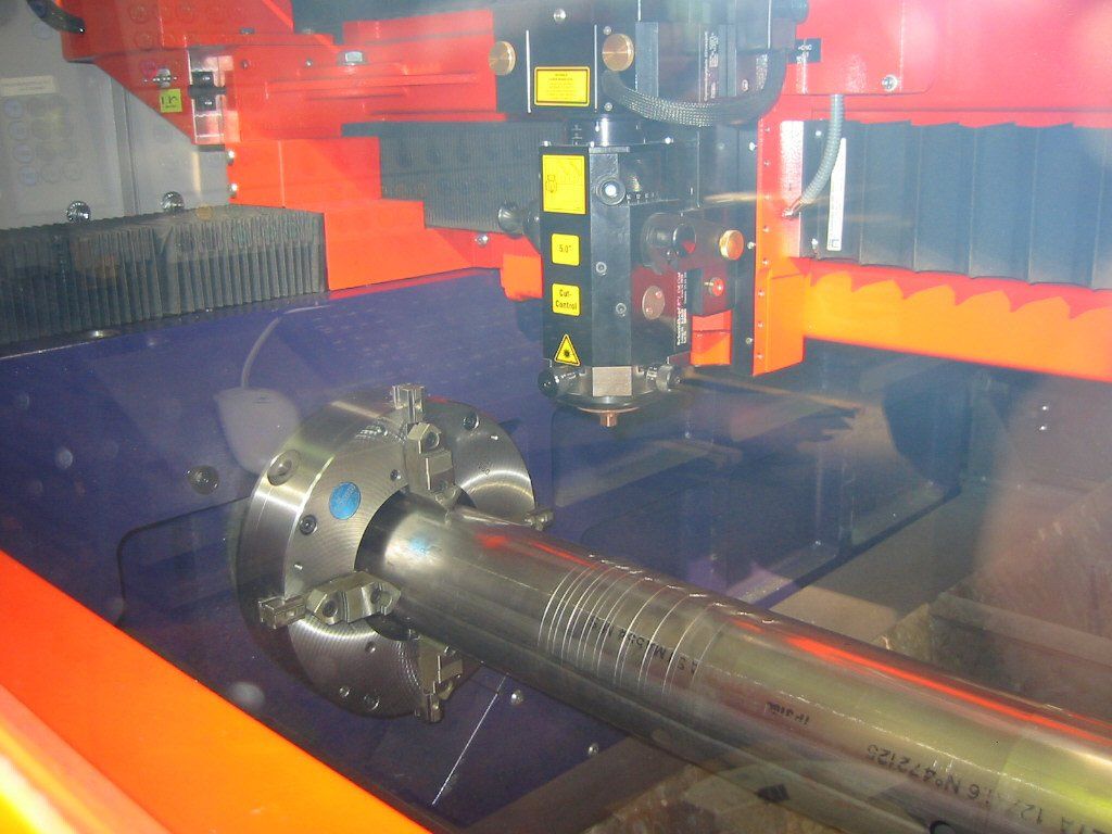 A laser cutter precisely cuts a metal cylinder held in a chuck. The machine is orange and black.