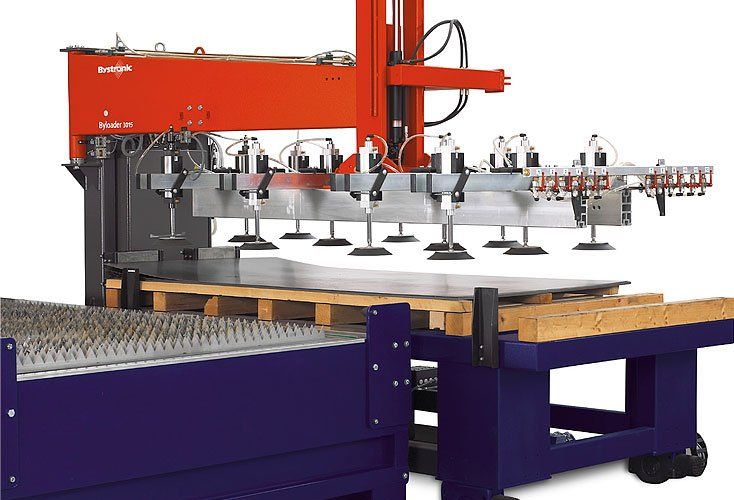 A robotic arm with suction cups lifts a metal sheet from a pallet in a factory setting. The machine is red, gray, and purple.