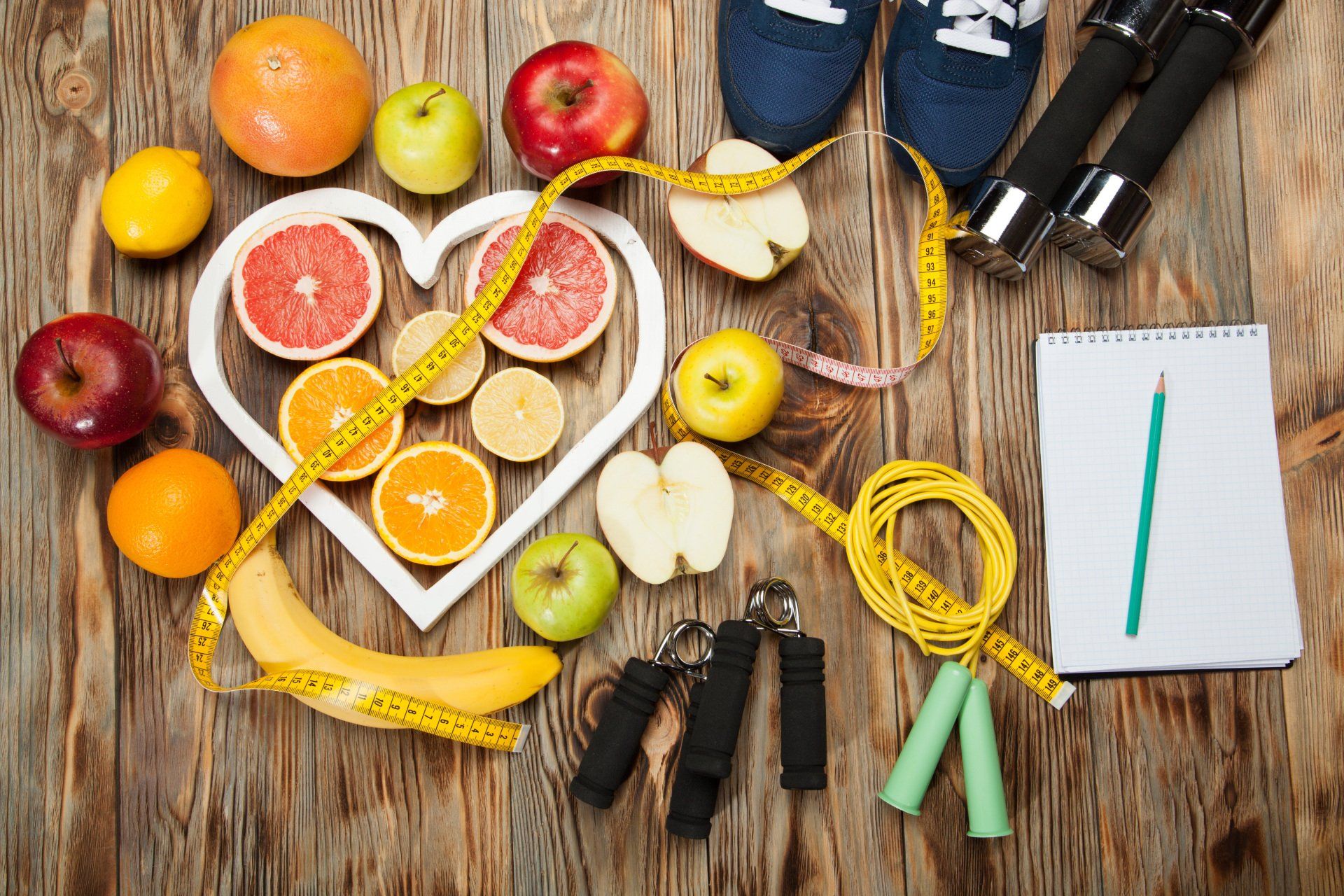 Una mesa de madera cubierta con fruta, cinta métrica, cuerda para saltar, mancuernas y zapatillas.