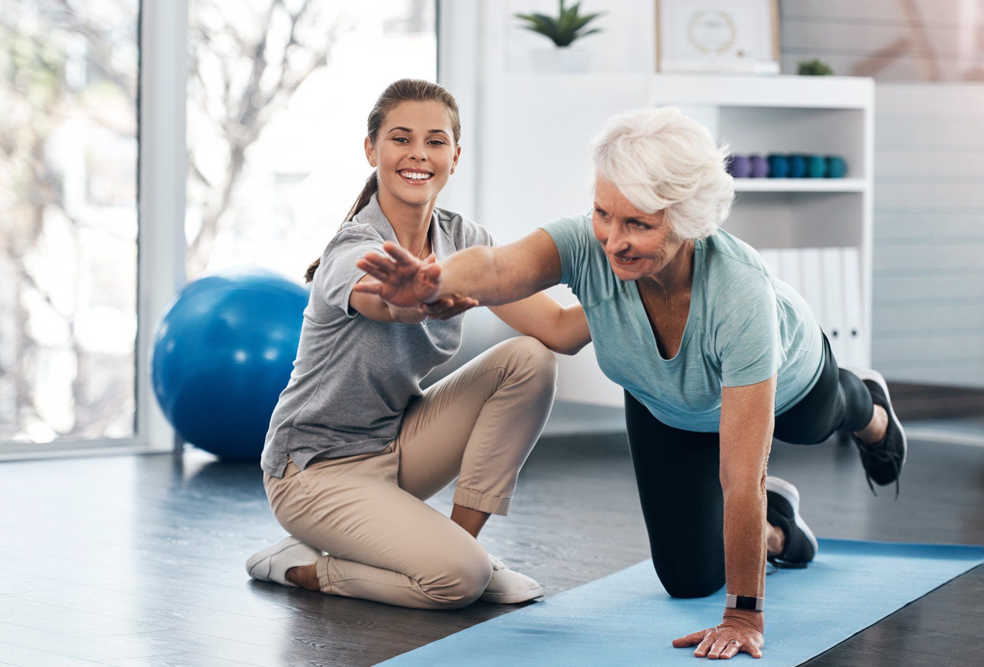 Woman assisting another woman with a balance exercise on a mat in a therapy room.