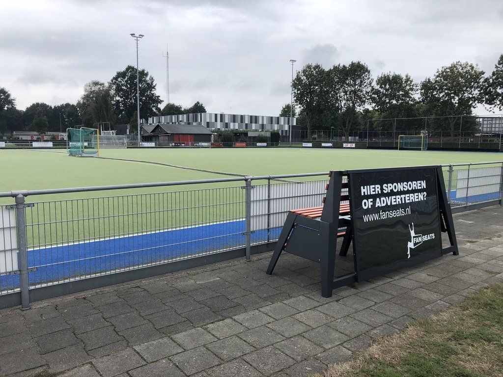 Een hockeyveld op een bewolkte dag. Op de voorgrond staat een Fanseats bank. De bank staat op een stenen pad.