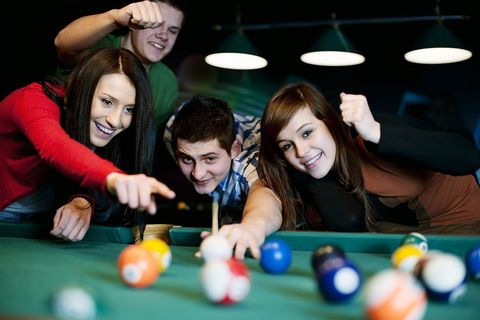 Friends playing pool, cheering, pointing, under bright lights.