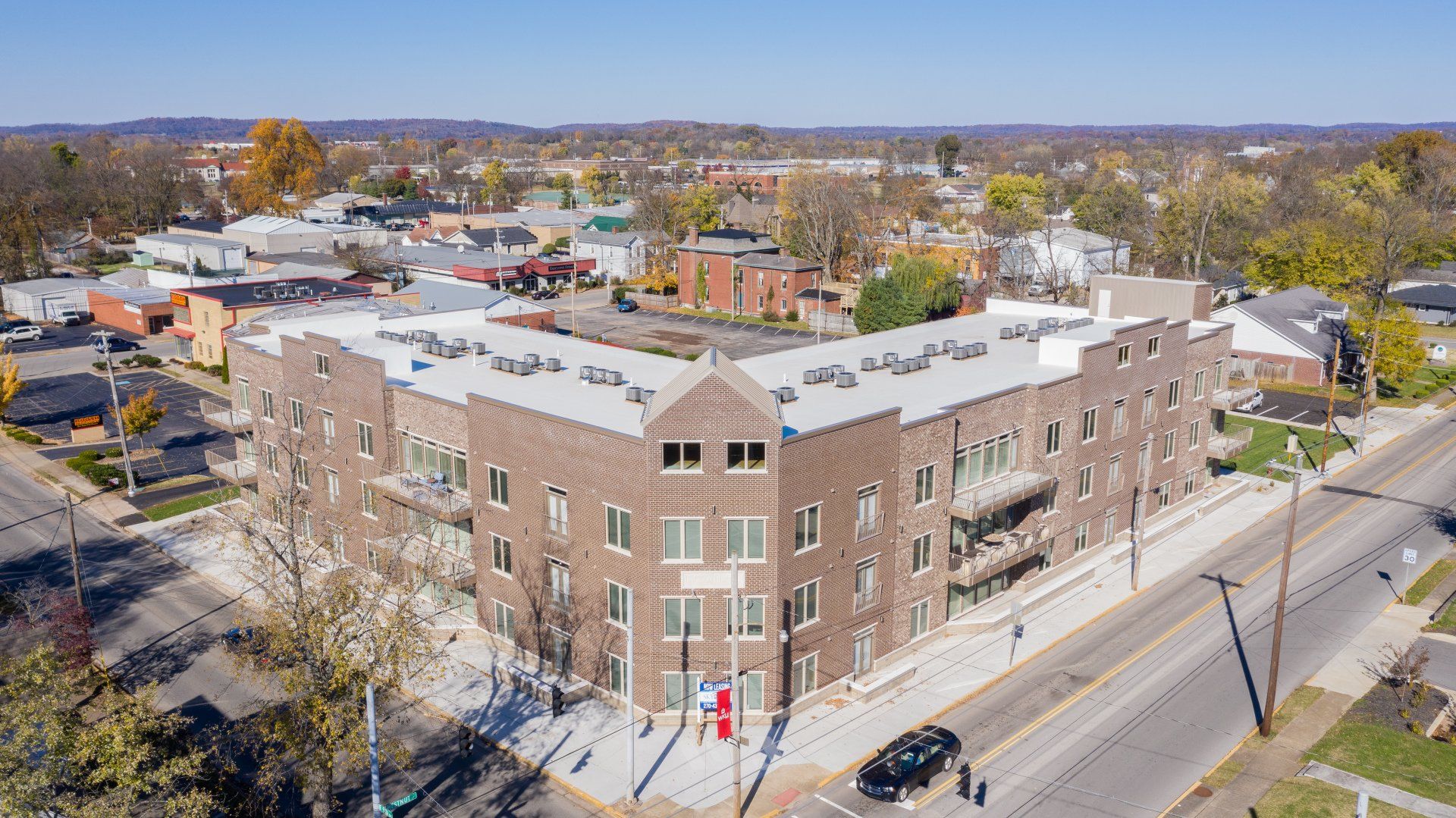 Brick apartment building on a corner lot with cars parked nearby, trees, and buildings in the background.