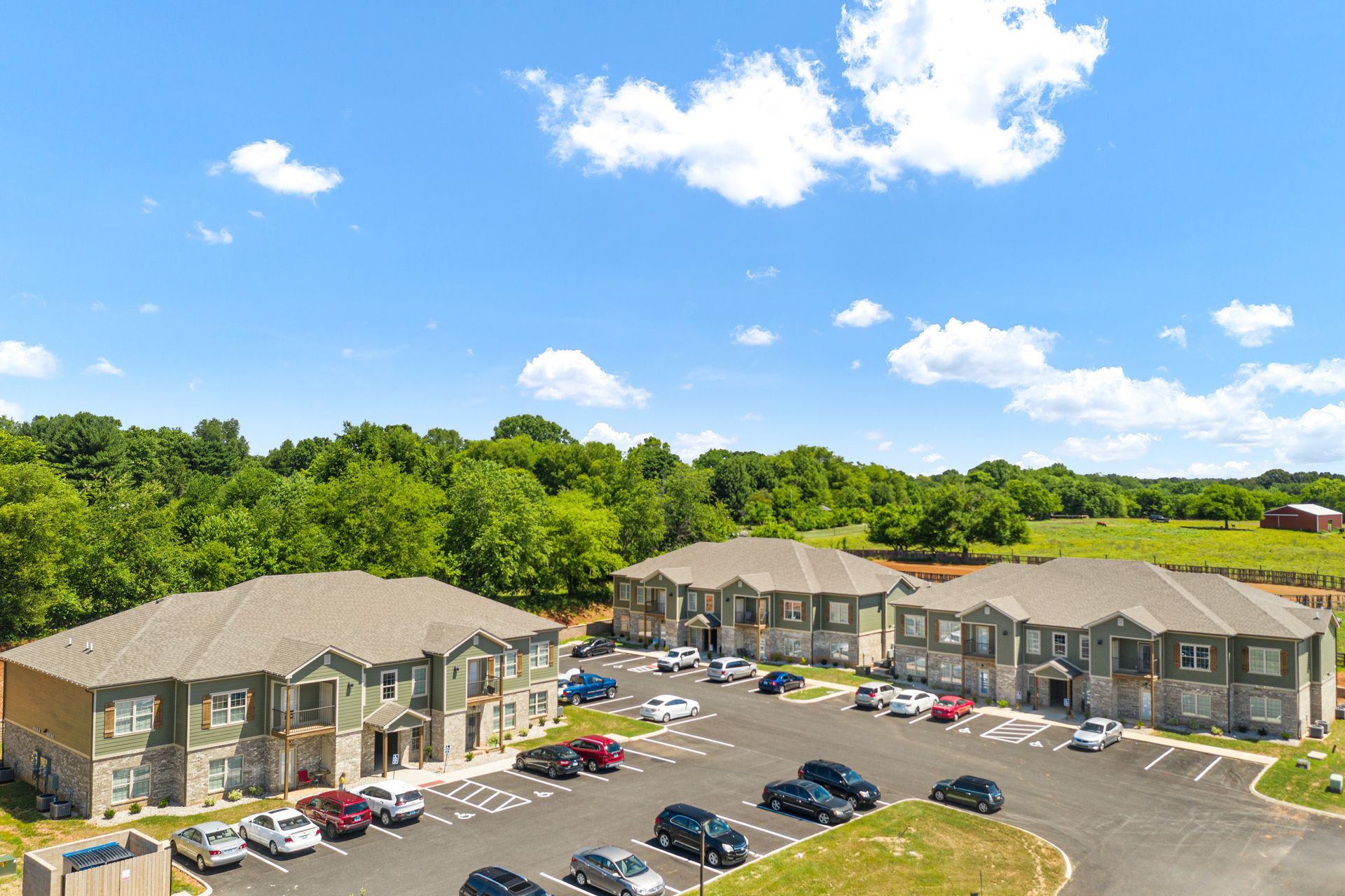 Apartment complex with gray buildings, parking lot, cars, and surrounding trees under a blue sky.