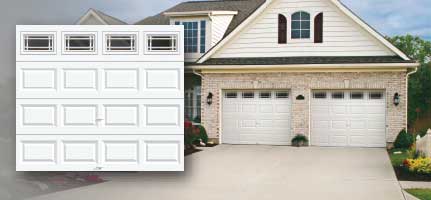A white garage door with rectangular windows, displayed next to a house with white garage doors and a concrete driveway.