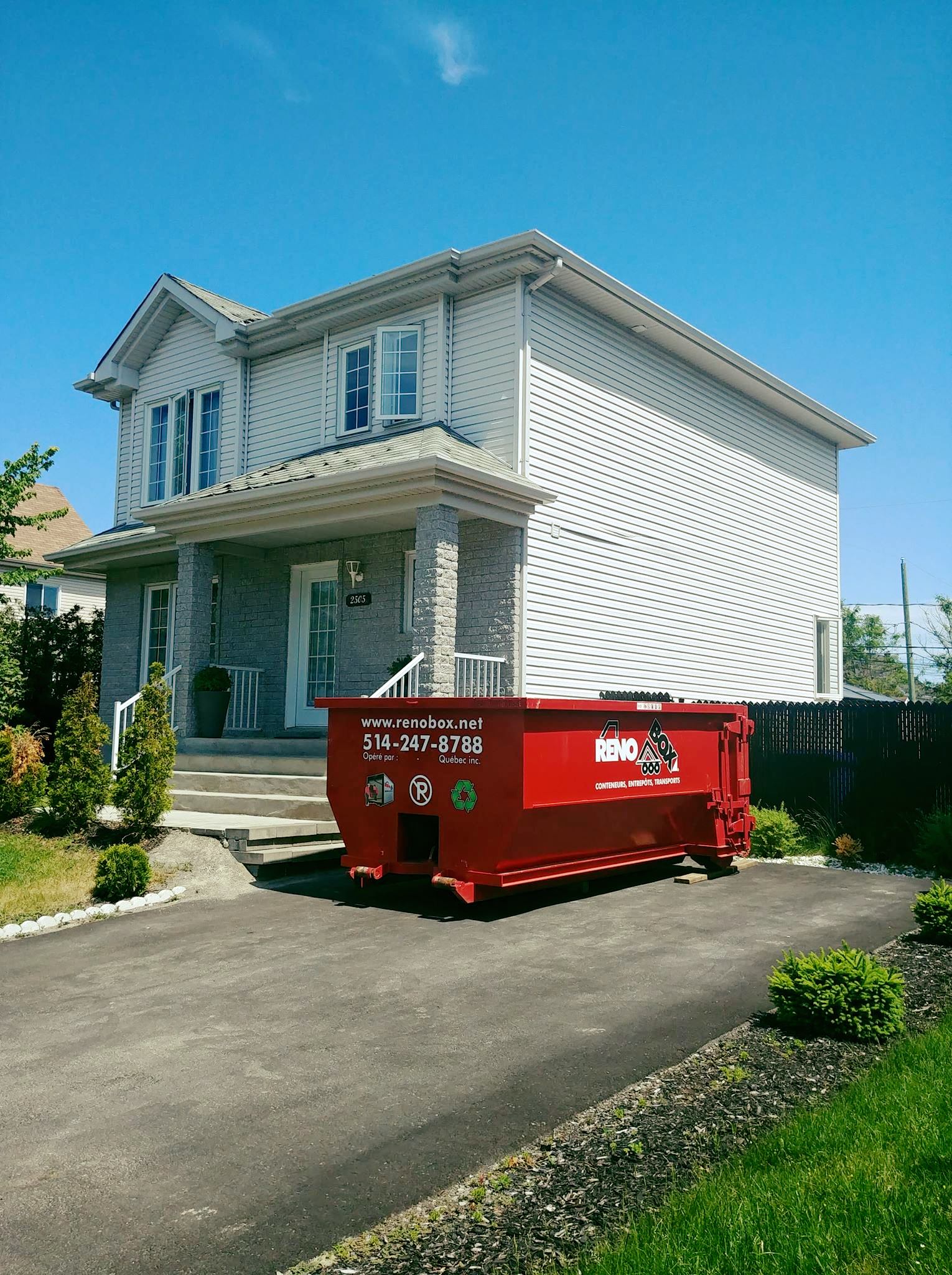 Une benne à ordures rouge se trouve devant une maison à deux étages avec une allée noire et une pelouse verte.