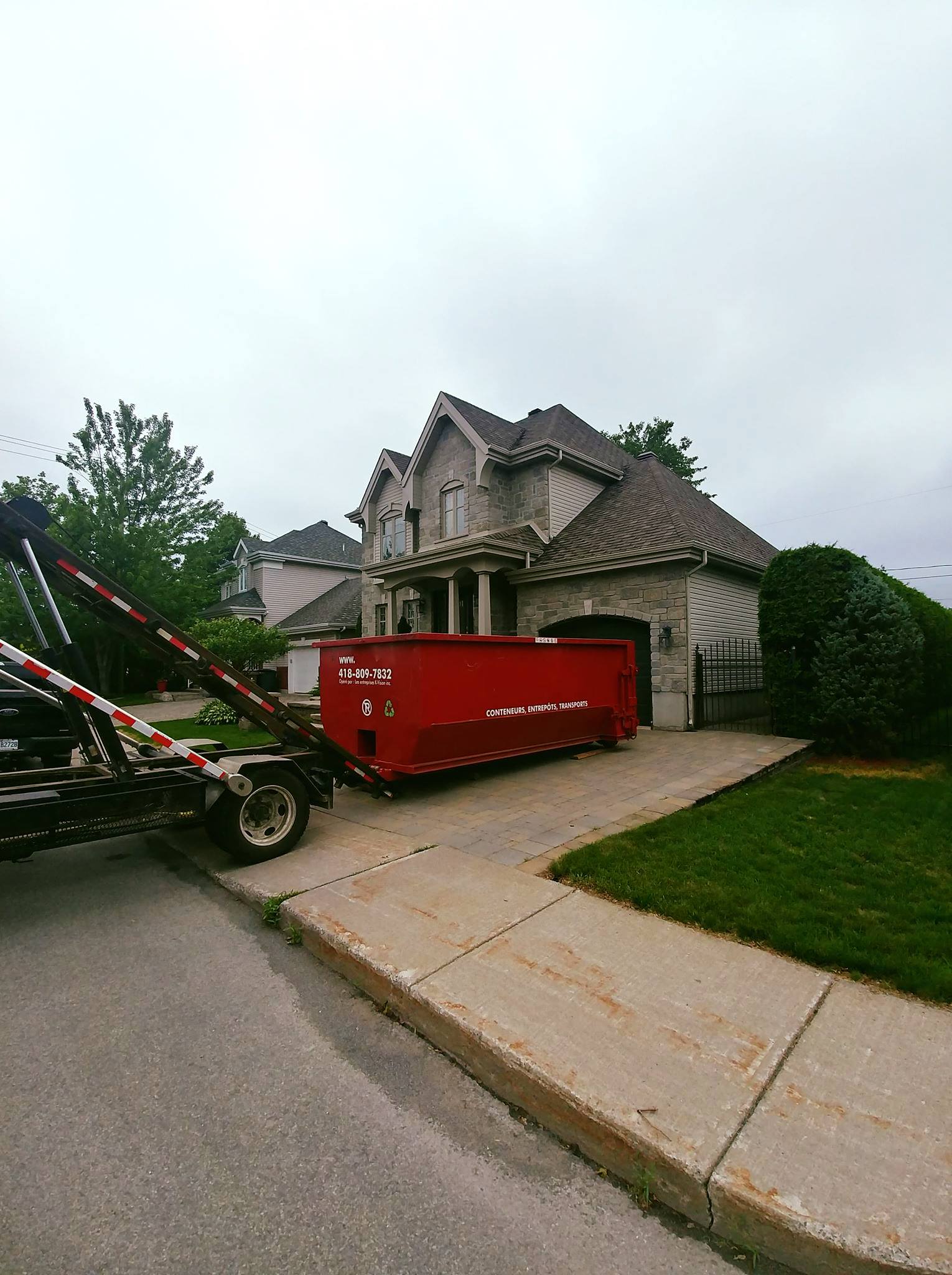Un camion décharge une benne à ordures rouge garée dans l'allée d'une maison en pierre.