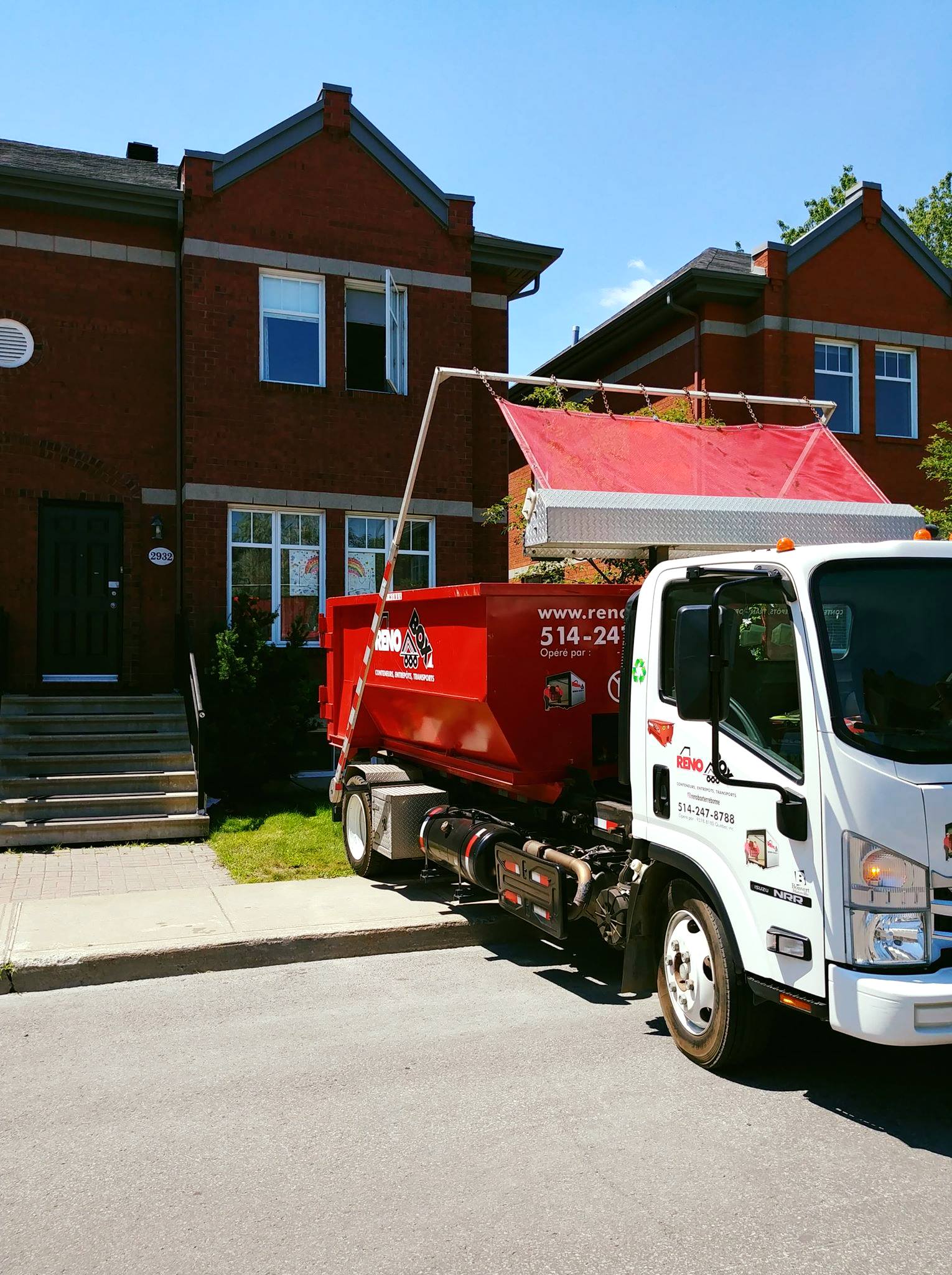 Un camion-benne rouge est stationné dans la rue devant un bâtiment en briques où des travaux de toiture sont en cours.