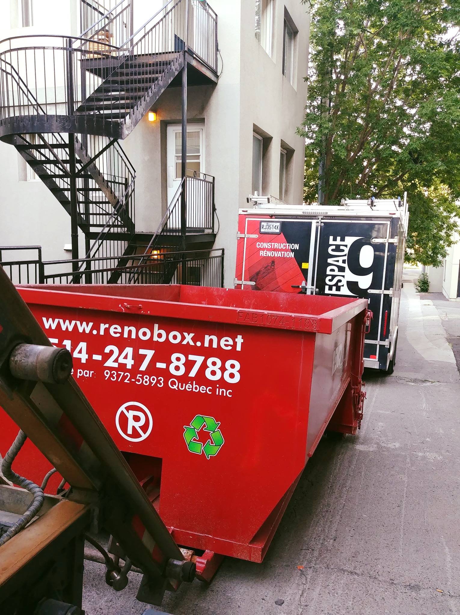 Une benne à ordures rouge dans une ruelle, avec un camion noir et blanc derrière. Un escalier métallique noir sur un bâtiment.