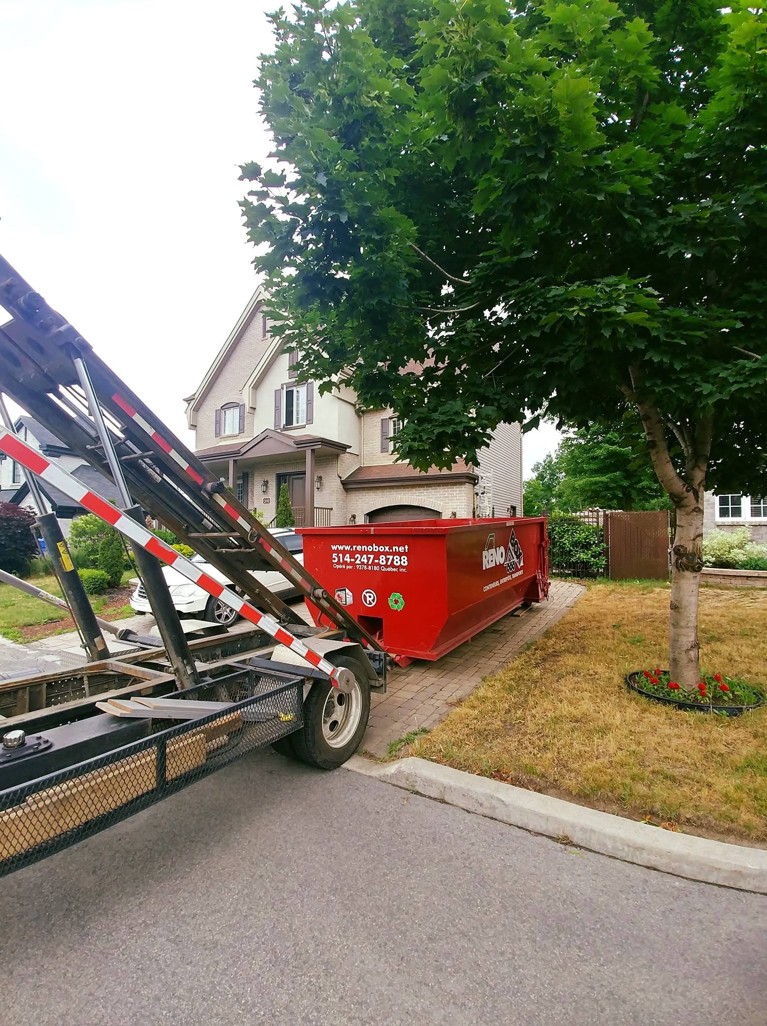 Une benne à ordures rouge est livrée sur une remorque dans une rue résidentielle, à côté d'une maison et d'un arbre.