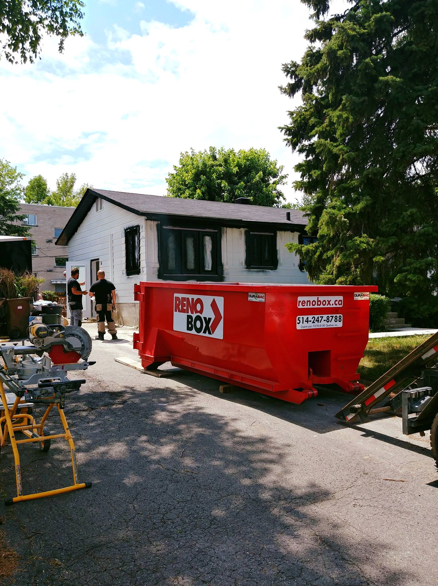 Une benne à ordures rouge devant une maison blanche, deux personnes à proximité. Outils de chantier visibles. Belle journée ensoleillée.
