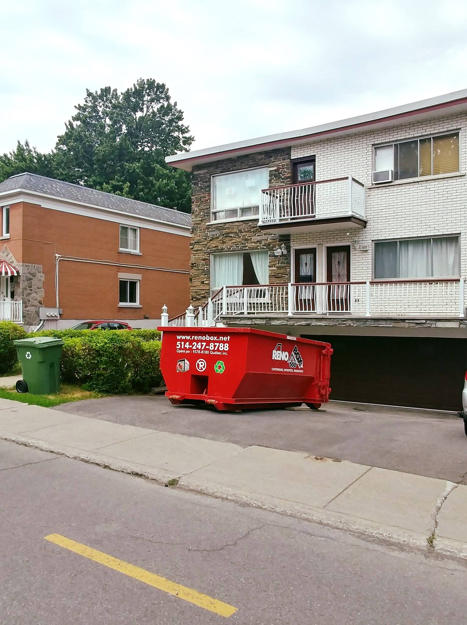 Une benne à ordures rouge se trouve devant un bâtiment de deux étages à façade en pierre, à côté d'une maison en briques et de buissons verts.