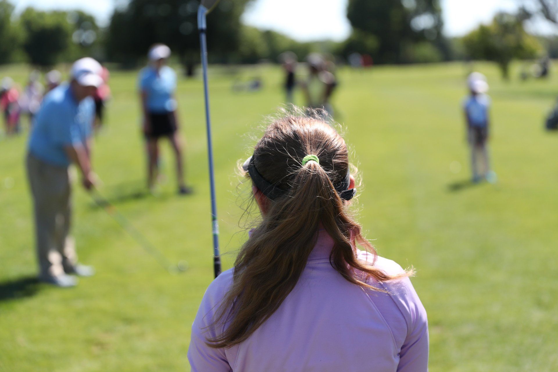 Woman with brown hair in ponytail watching golfers on a green field on a sunny day.