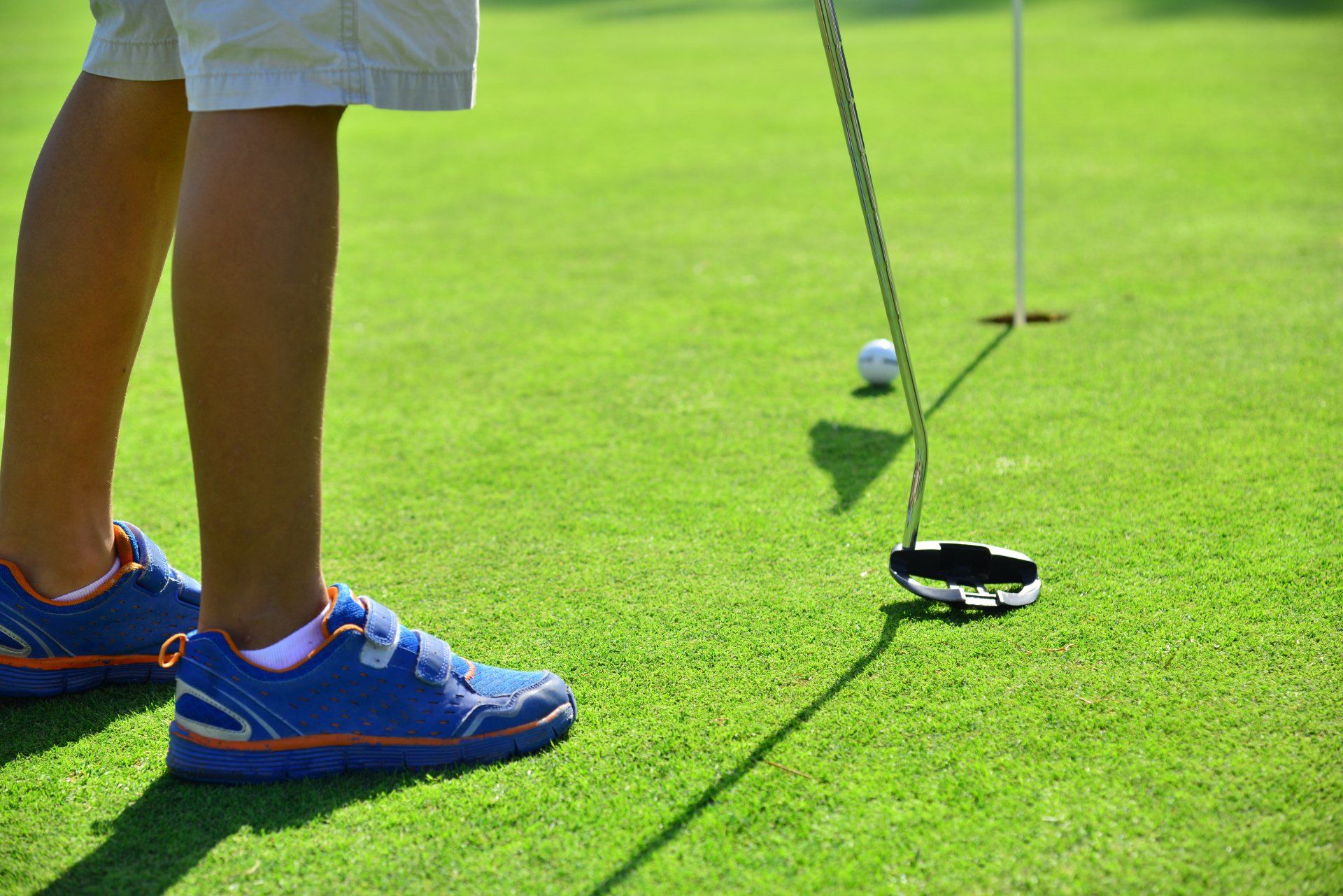 Person putting golf ball on green, blue shoes, white shorts. Flag in hole.