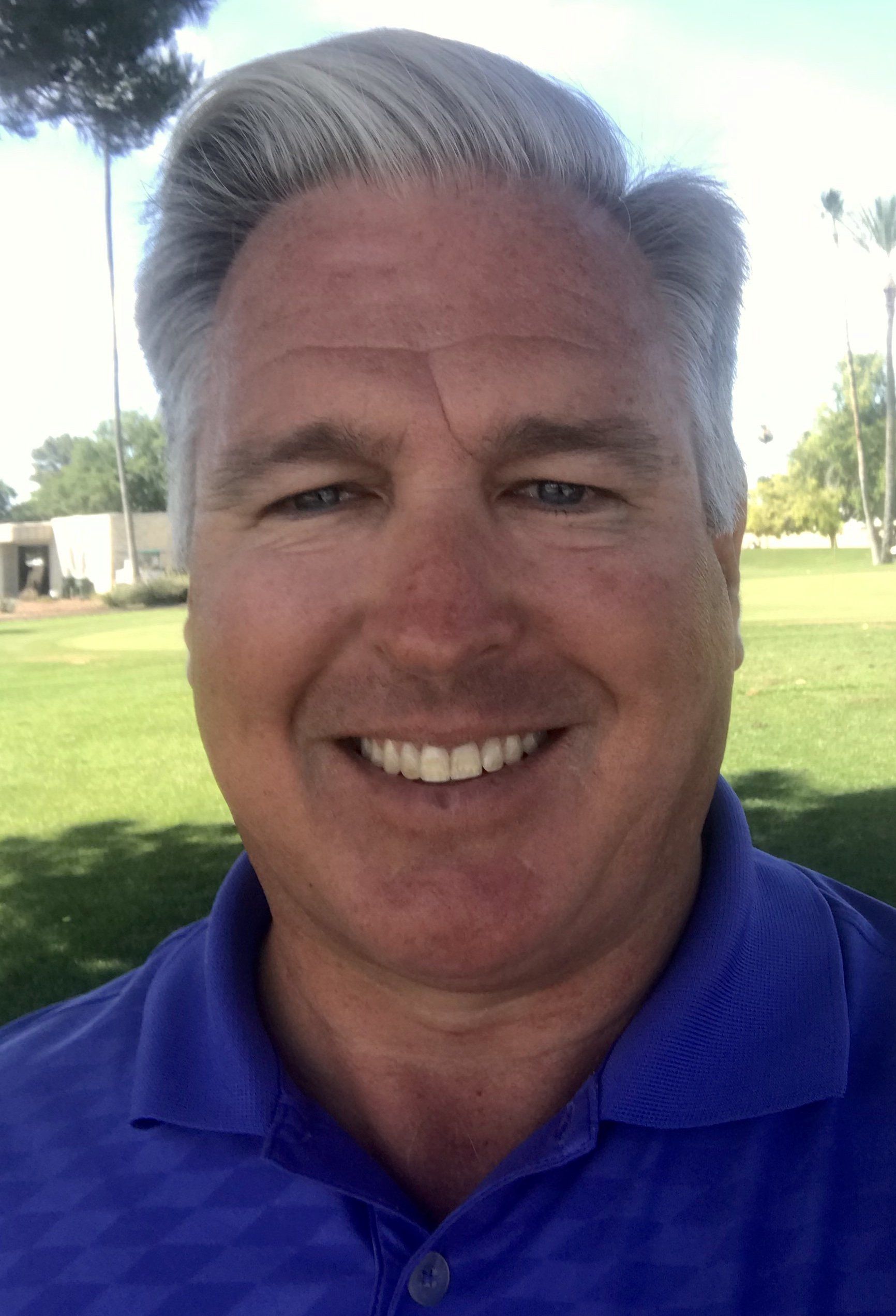 Man smiling outdoors in blue polo shirt. Green grass background.