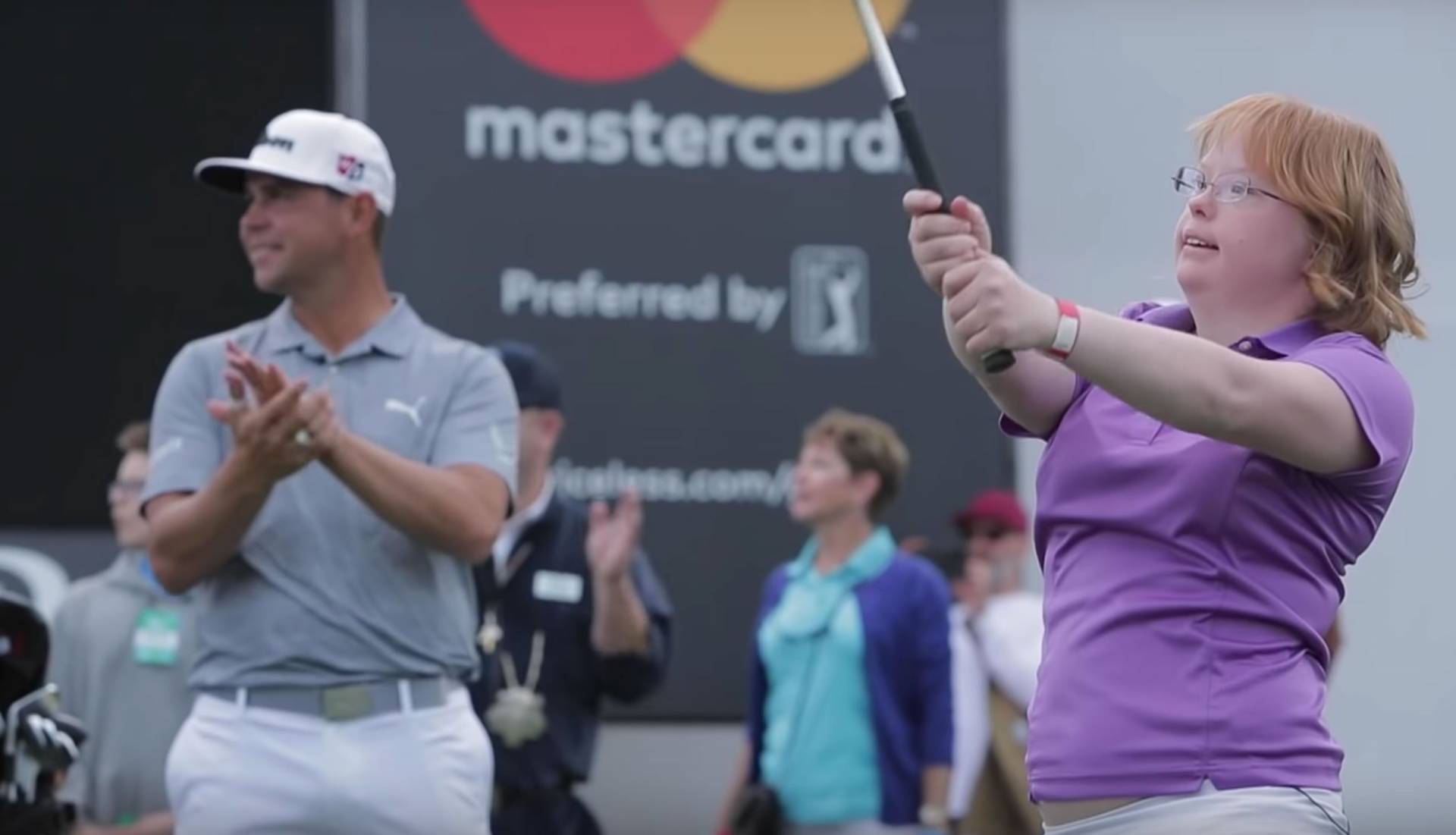 Golfer applauds girl with Down syndrome swinging a club, at a golf tournament with a Mastercard sign.