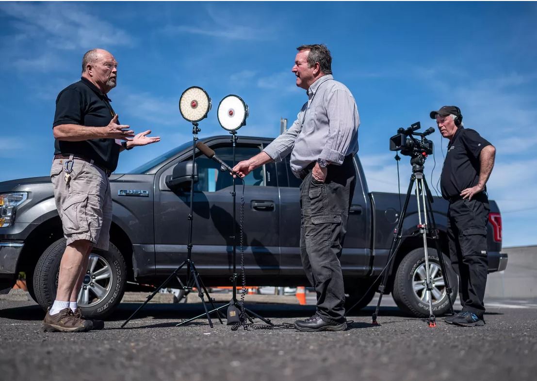Two reports interviewing a man. They are all standing in front of a dark silver truck