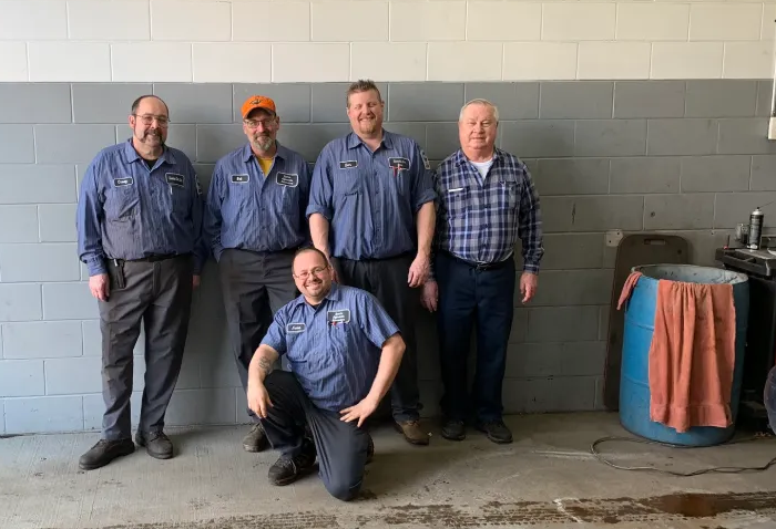 Five auto mechanics in blue work shirts pose in a garage. One kneels. Grey wall.