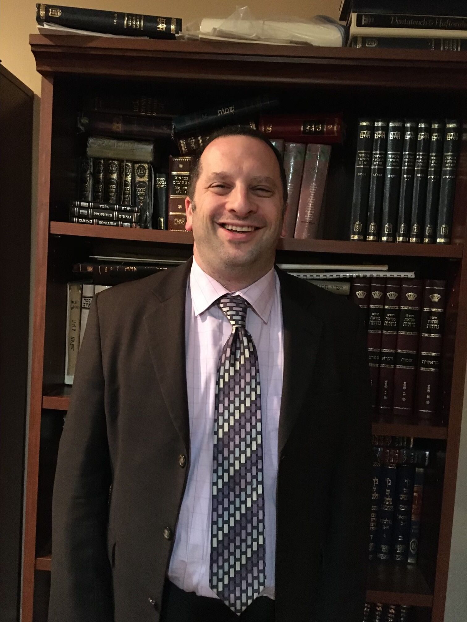 Man in a suit smiling in front of a bookshelf-lined office cabinet
