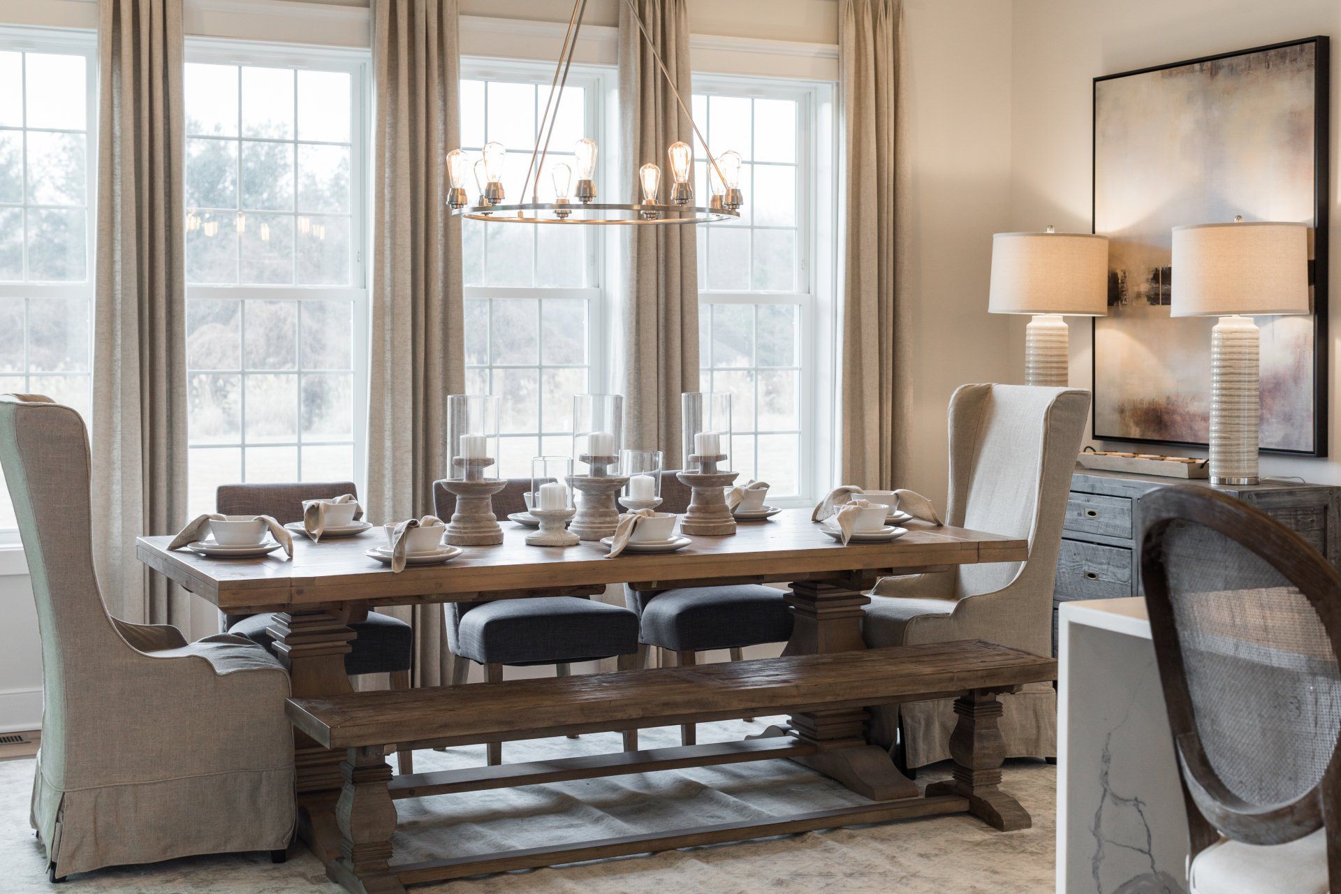 Elegant dining room with a wooden table, bench, and chairs near large windows, with candles and a chandelier.