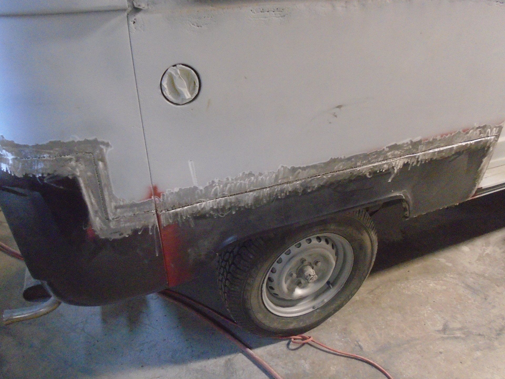 Car body panel repair. Gray car side with fuel door, black lower panel. Shows welding and grinding work.