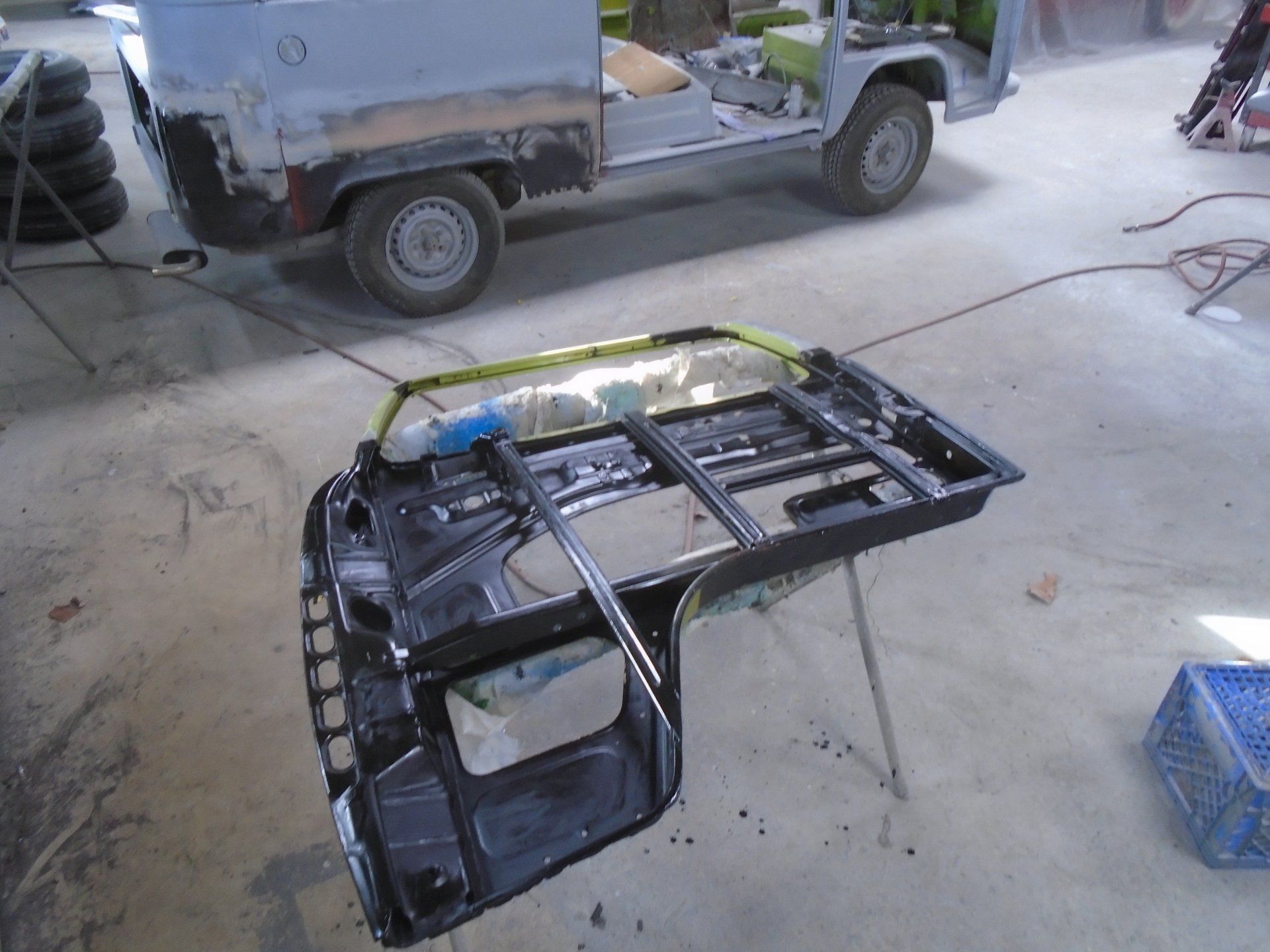 Black car frame on stands, with a light green Jeep truck in the background. Workshop setting.