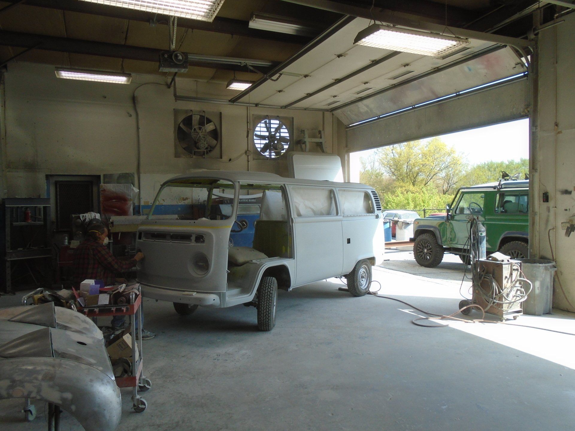 Inside auto body shop: partially restored VW van, green Land Rover visible, person working.