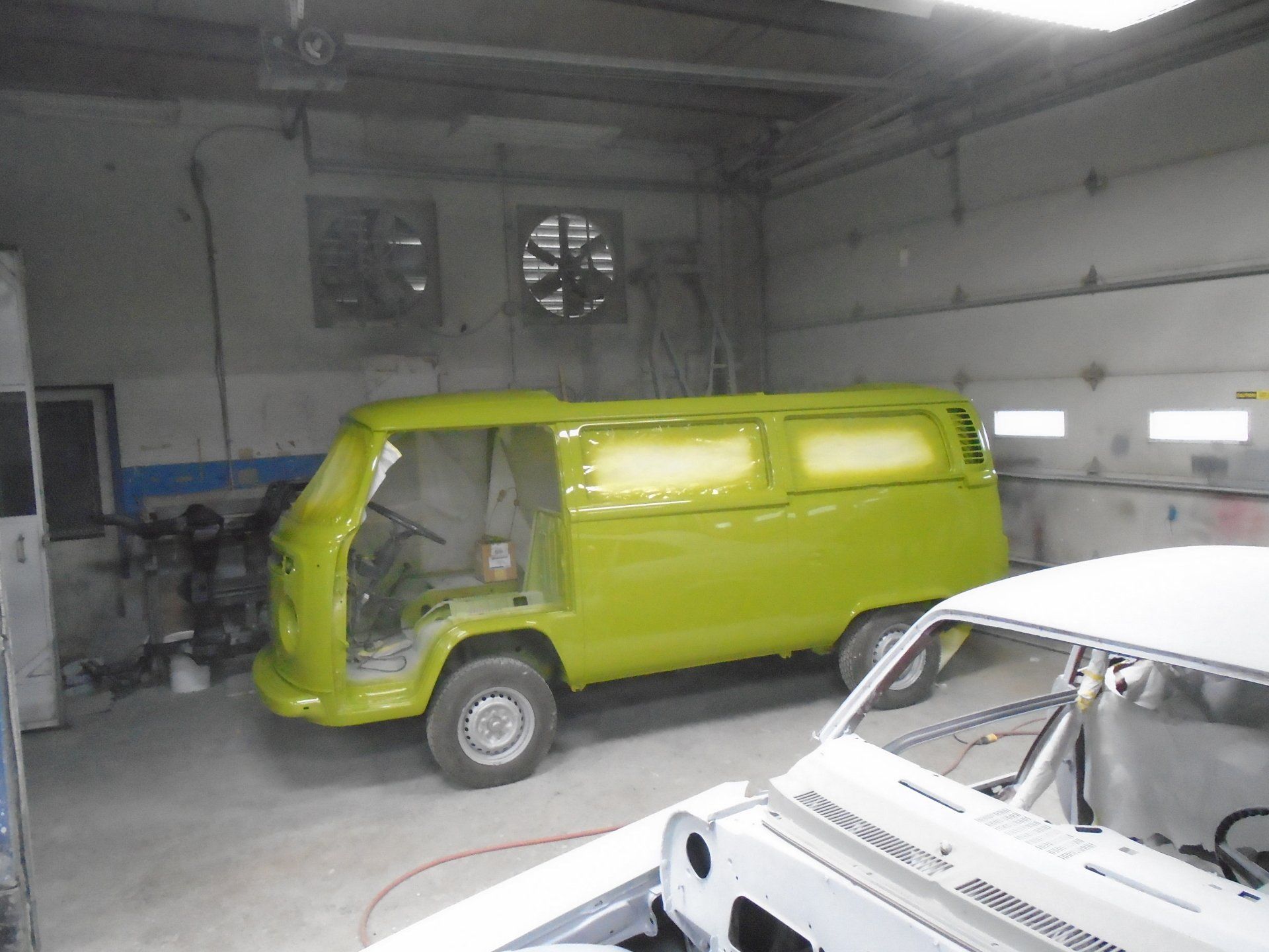 A lime green van in a workshop, partially painted. Another white car sits nearby.