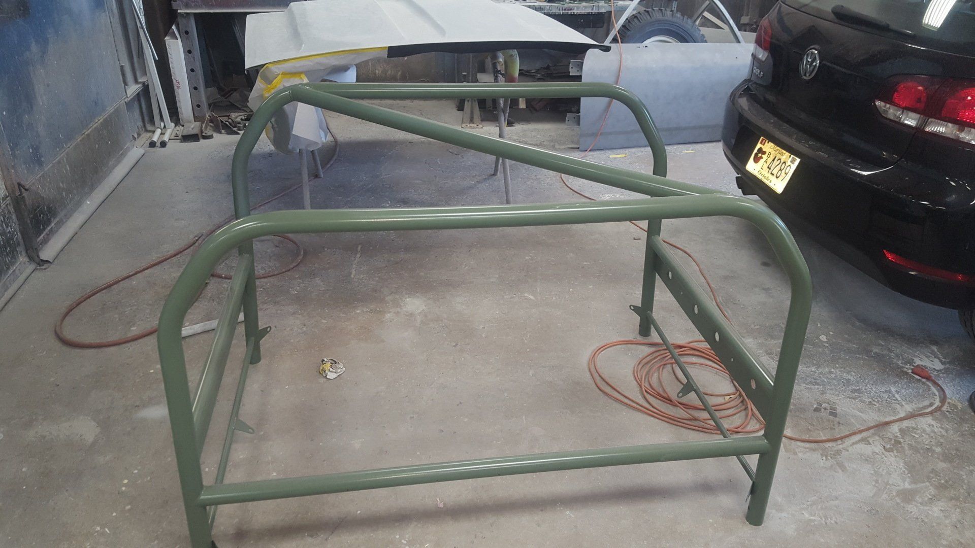 Green metal roll cage in a workshop, next to a black car.