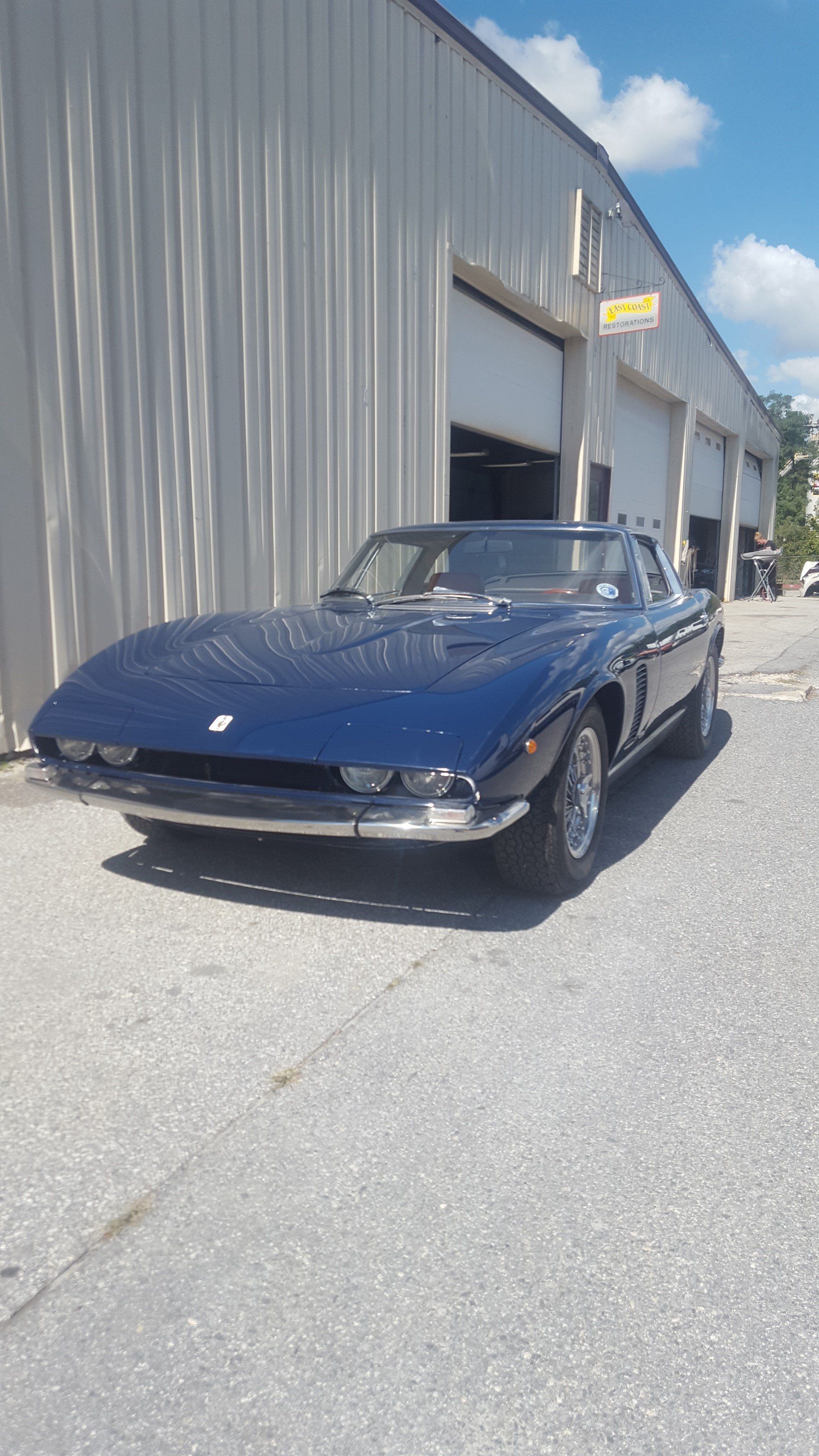 Blue Jensen Interceptor car parked in front of a gray industrial building on a sunny day.
