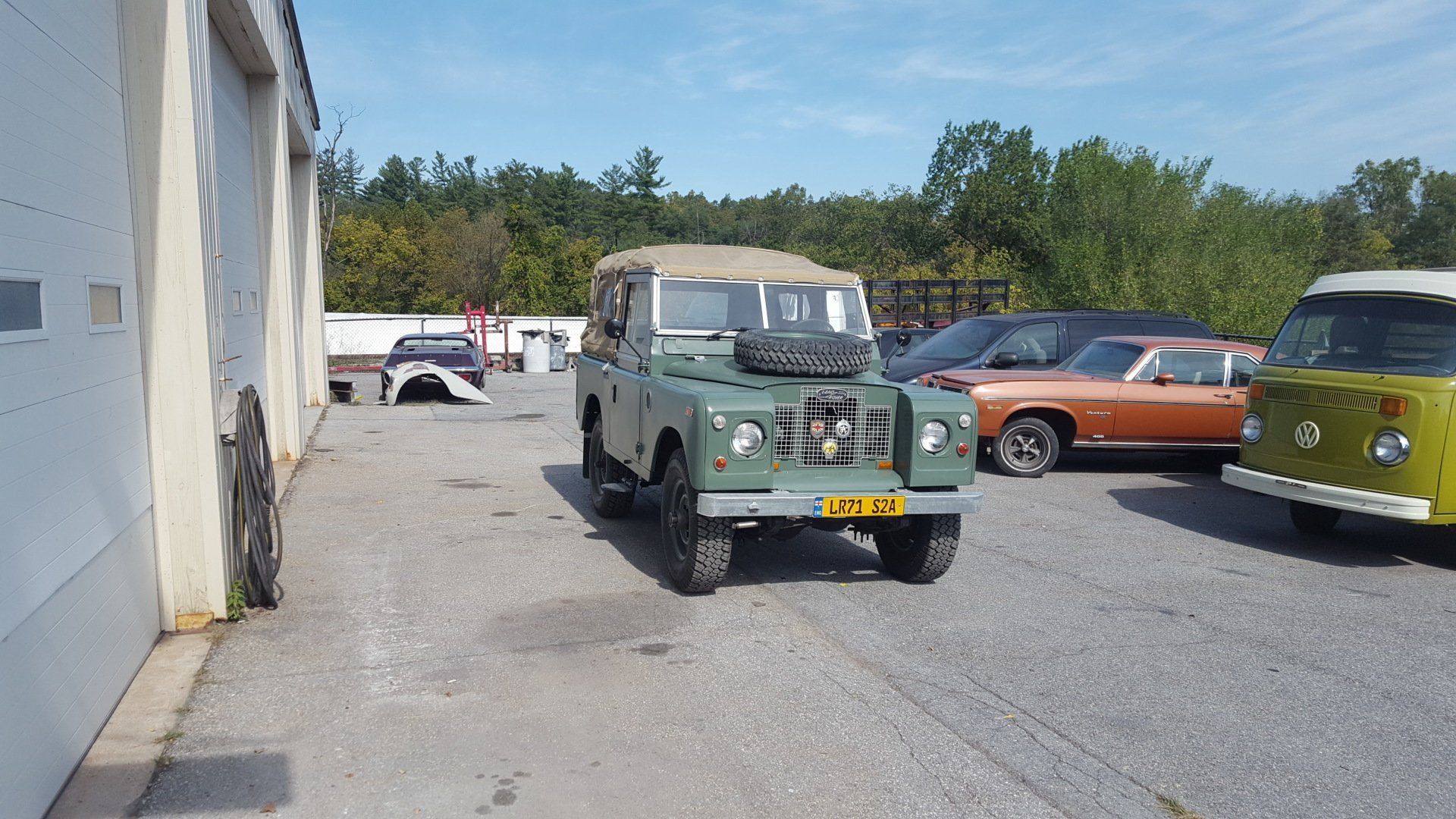 Green vintage Land Rover parked outside a building. Other cars are nearby, under a blue sky.