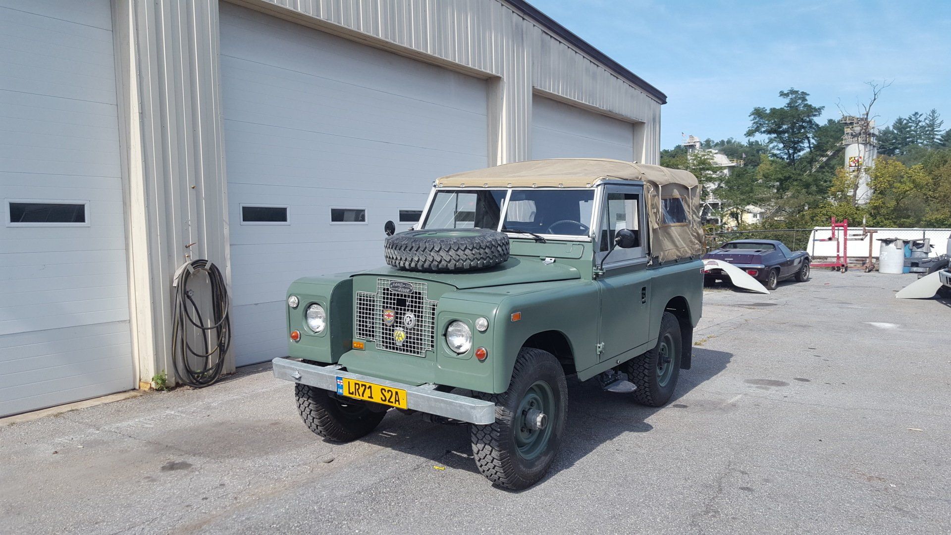 Green Land Rover parked in front of a garage, spare tire on the hood, tan convertible top.