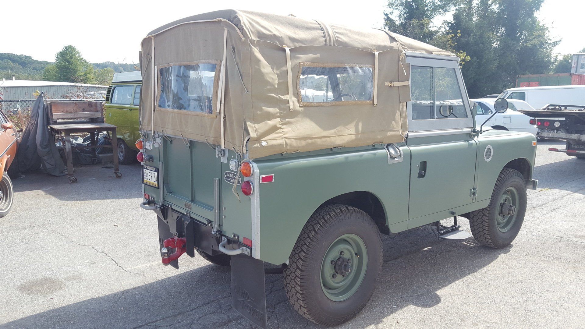 Green Land Rover Series 3 with tan canvas top. Parked on a paved area, trees in the background.