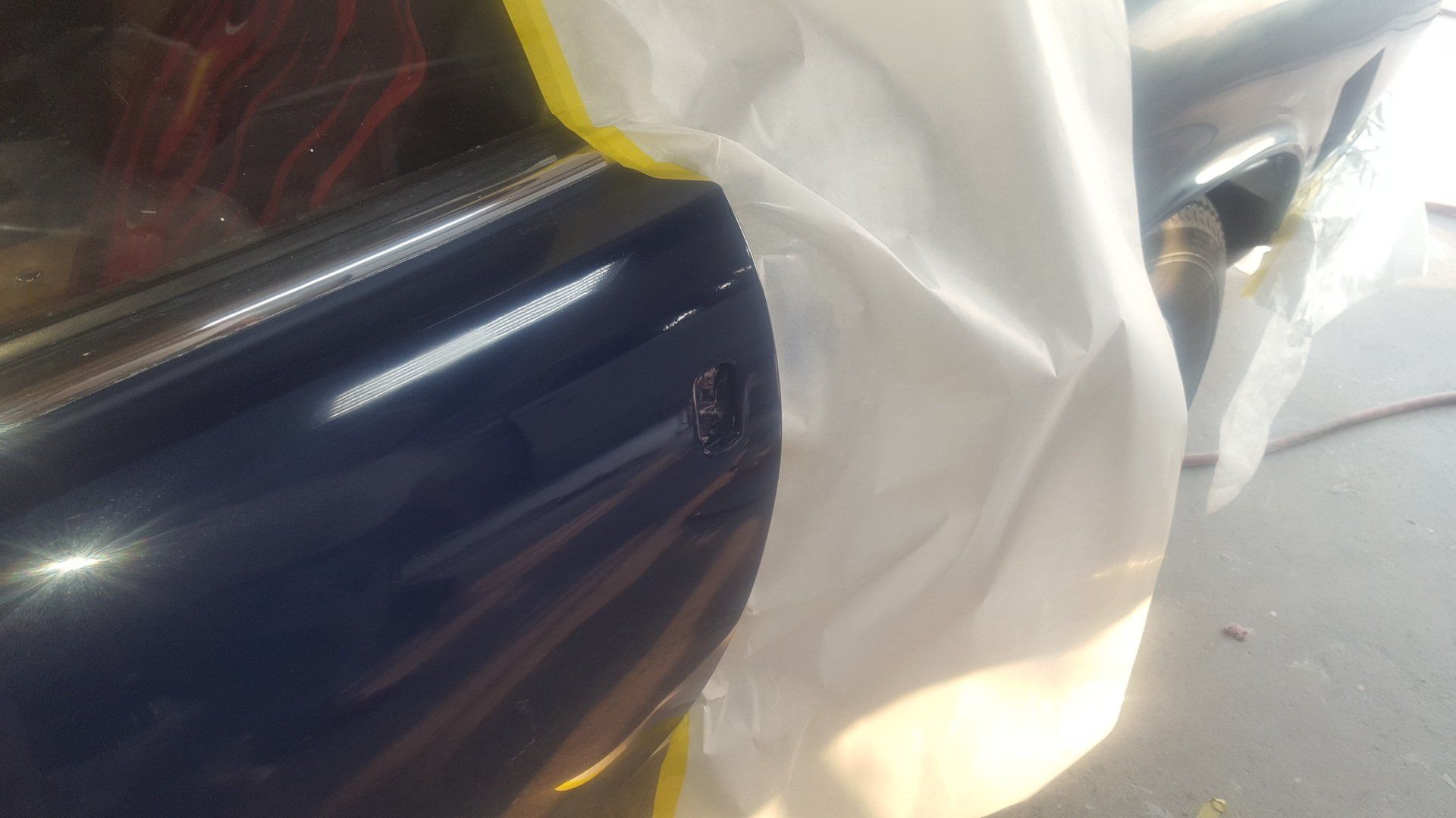 Dark blue car door being prepped for paint, covered in white plastic sheeting with yellow tape.
