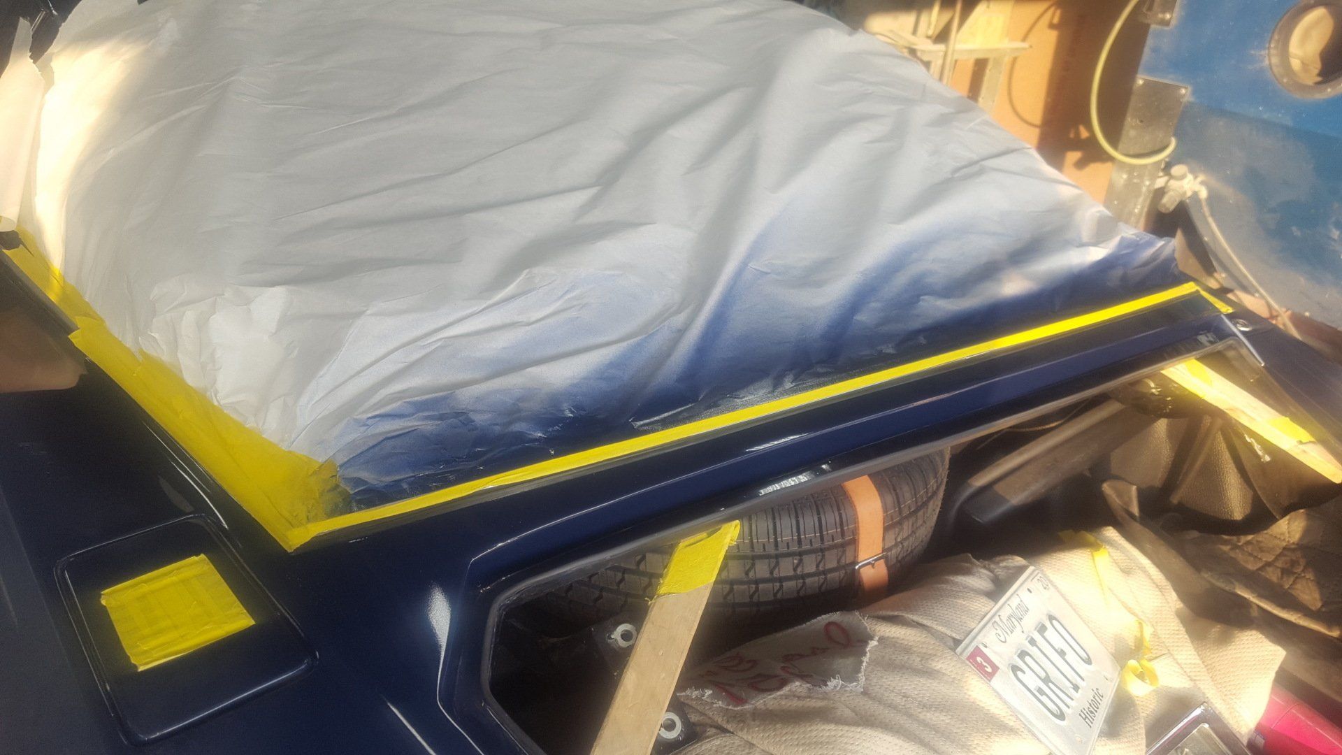 A car's painted blue body panel with yellow tape and a white tarp, in a garage.