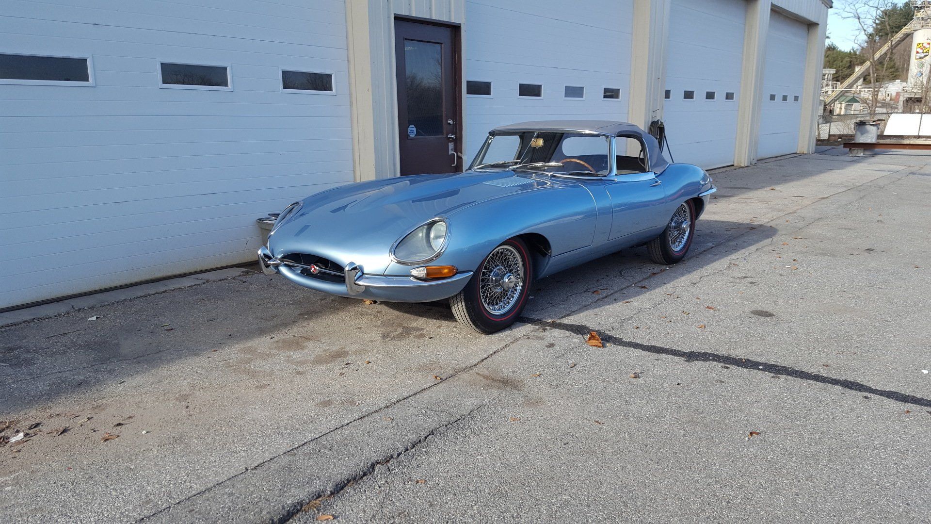 Blue Jaguar E-Type coupe parked outside a light blue building with a black door and garage doors.