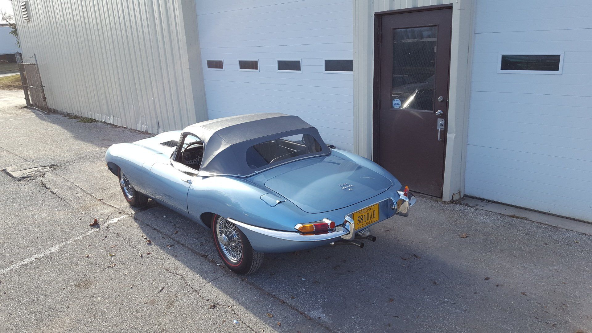 Blue Jaguar E-Type convertible parked in front of a white building with a brown door.