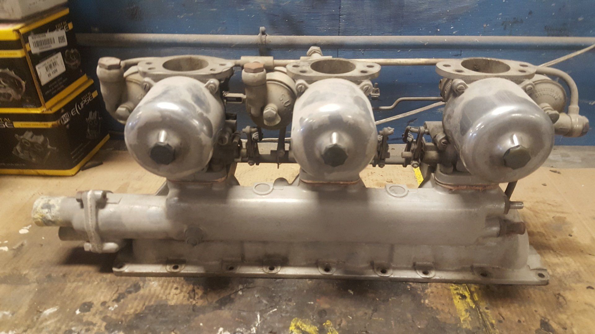 Three-carburetor intake manifold assembly, painted gray, resting on a wooden surface.