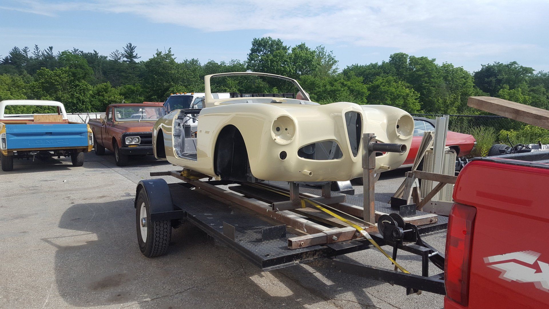 A classic convertible car body, painted beige, on a trailer. Other vintage vehicles are in the background.