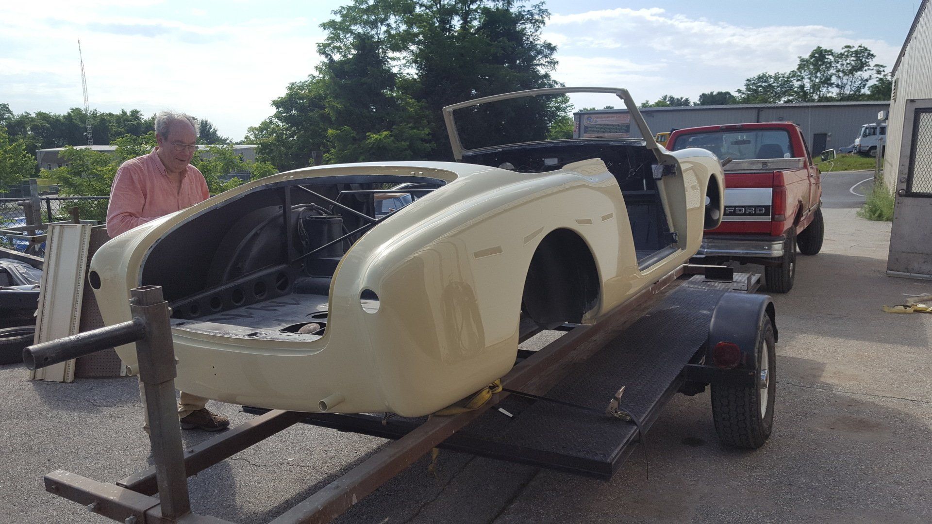 A car body on a trailer, cream-colored with an open cockpit, and a man near it outside.