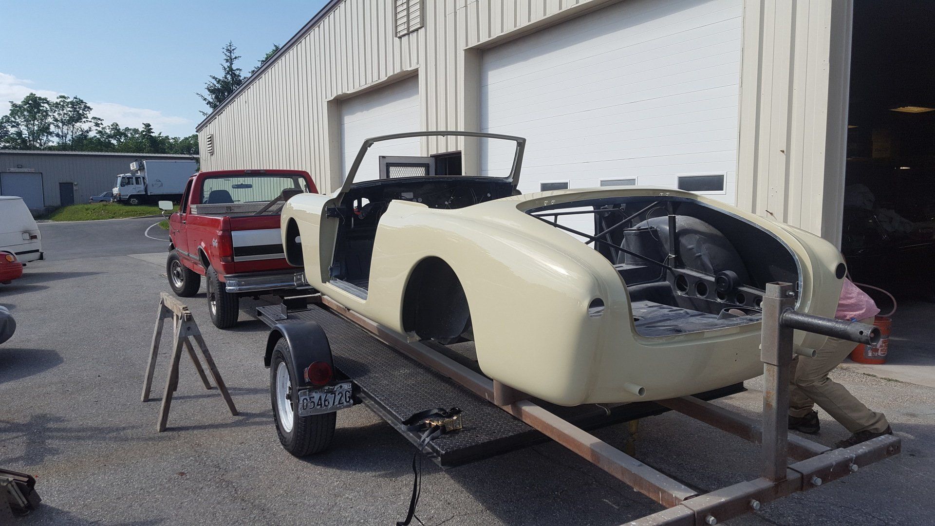 A cream-colored vintage convertible on a trailer in front of a garage, being prepped for restoration.