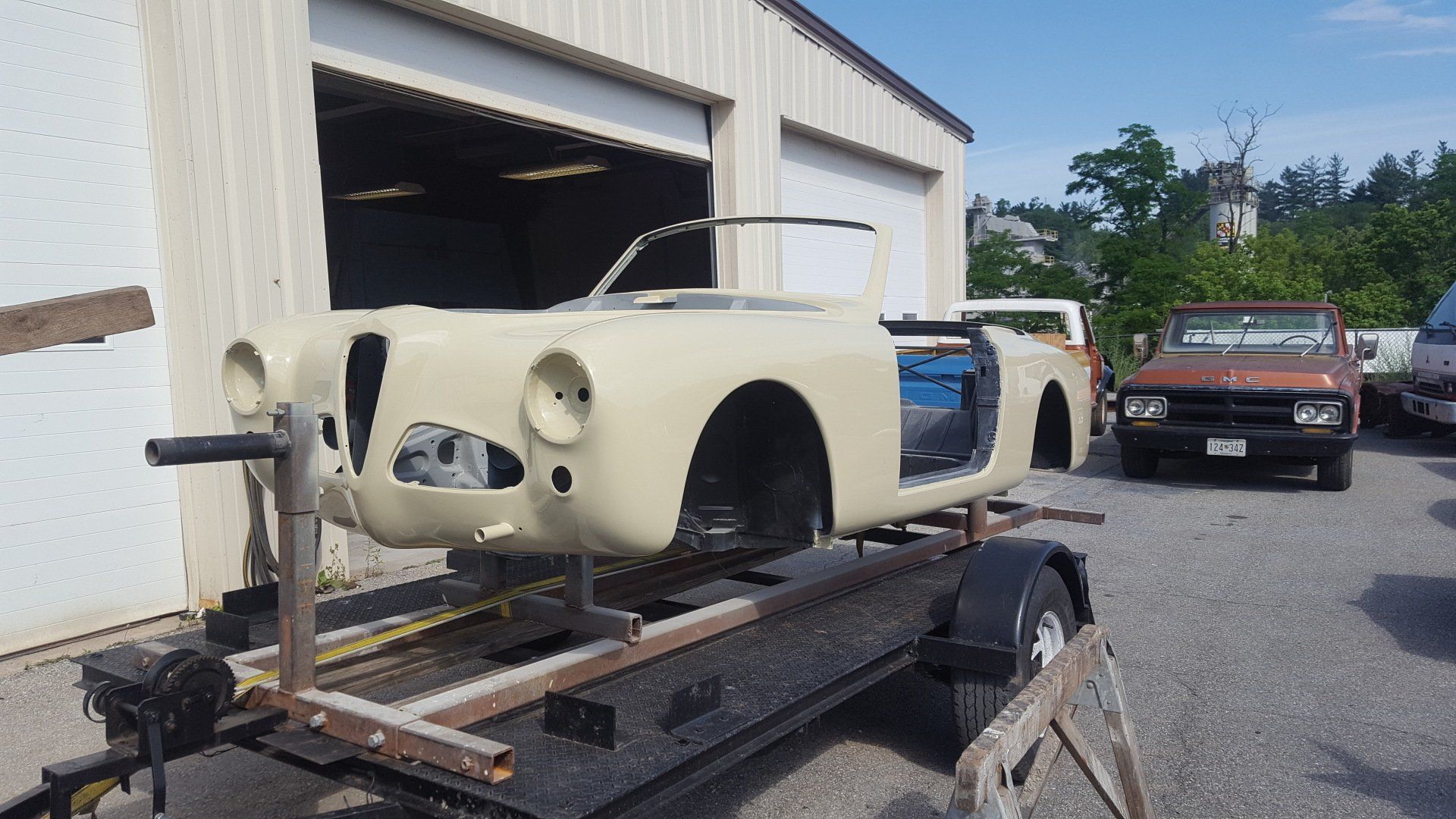 Beige car body on a trailer outside a garage; another car and building visible.
