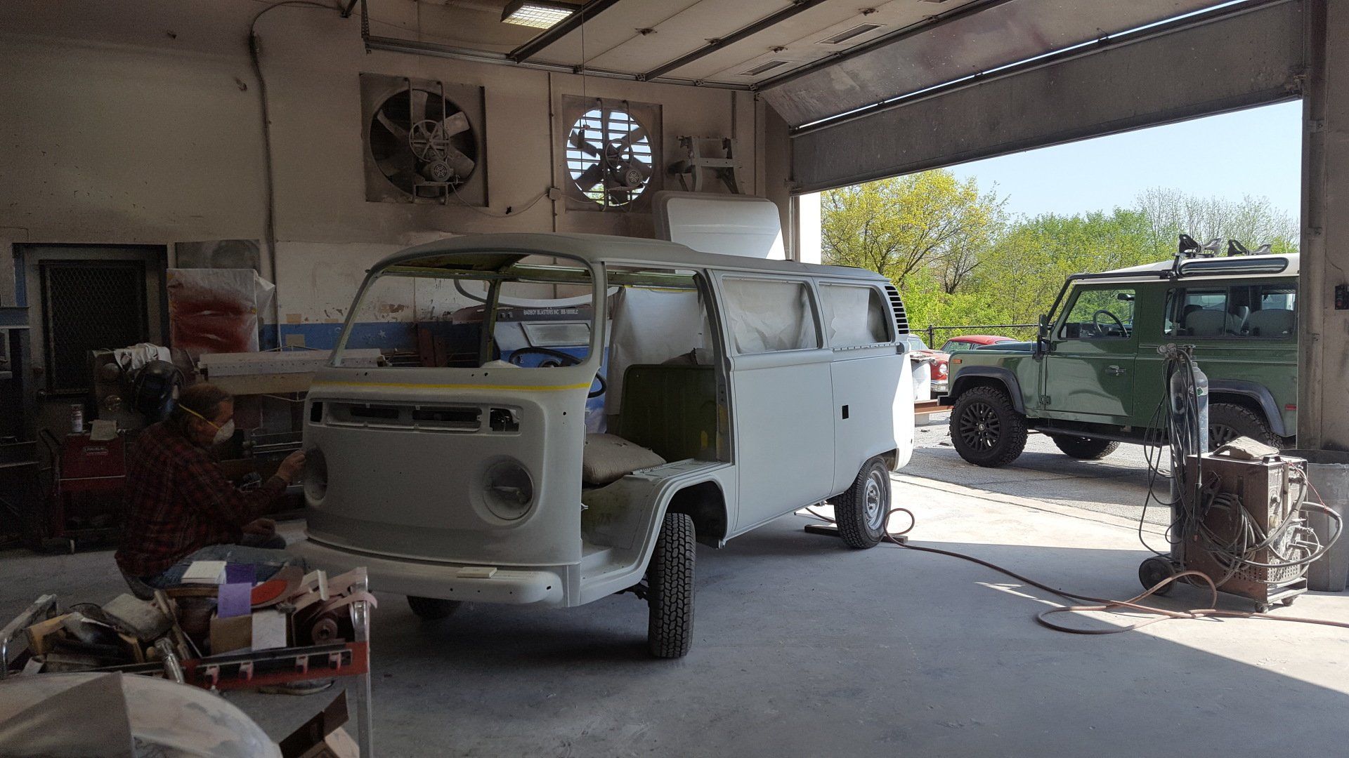 A light-colored VW van in a shop being worked on, a green SUV sits to the right, and a person welds on the left.