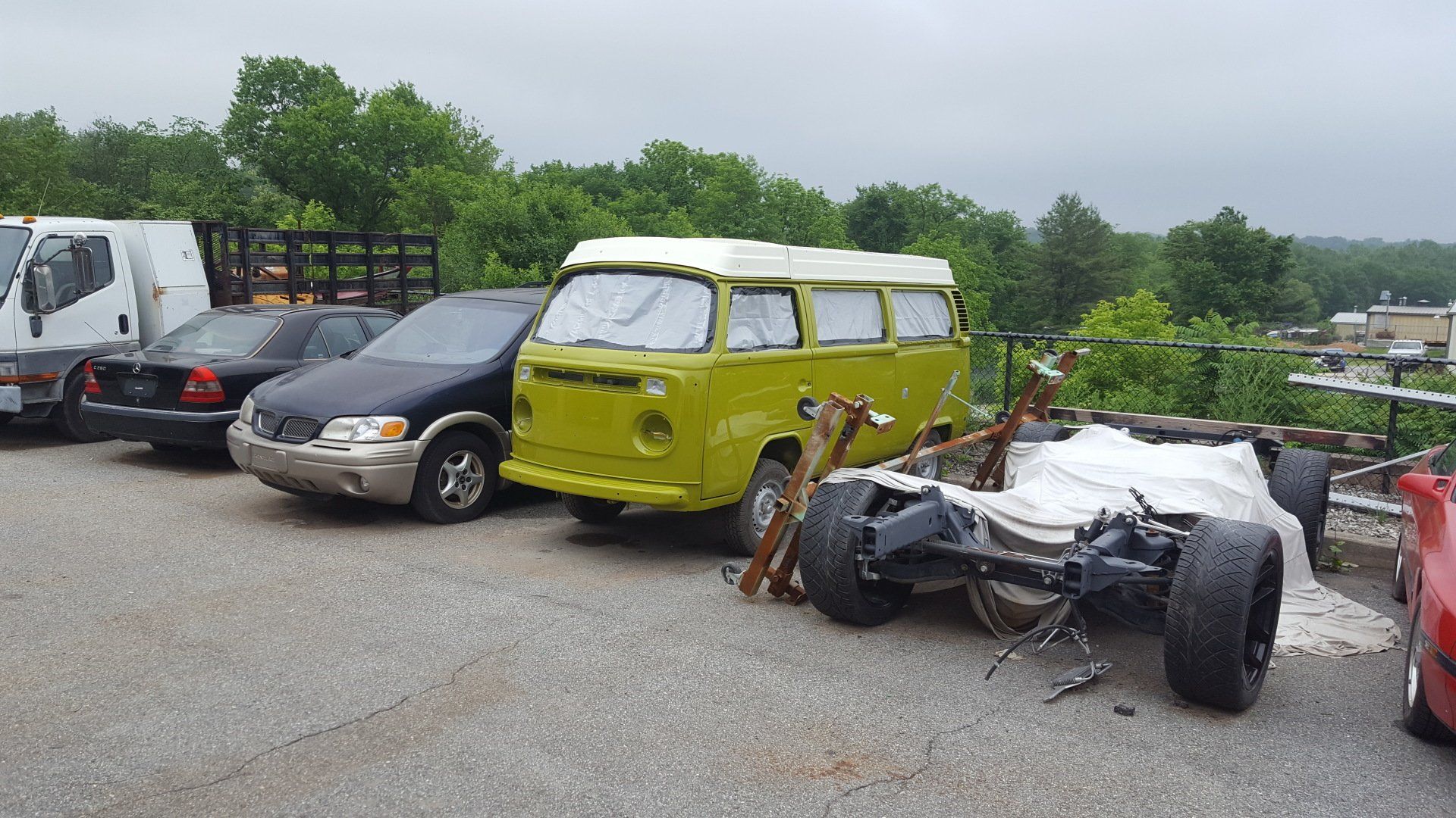 Yellow VW bus, other vehicles, and a dismantled car frame in an outdoor parking area.