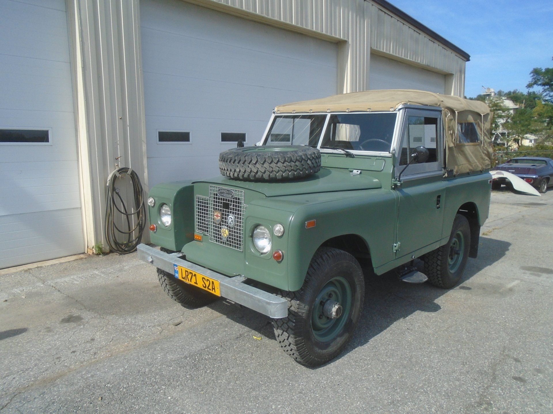 Green vintage Land Rover with a tan soft top, parked in front of a building. Spare tire on the hood.