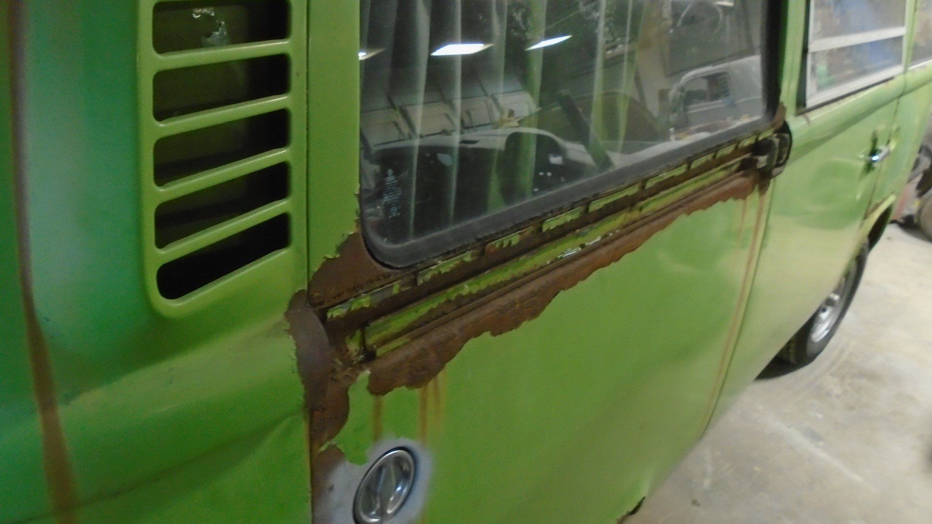 Green vintage VW bus side, showing extensive rust around the door and window frame.