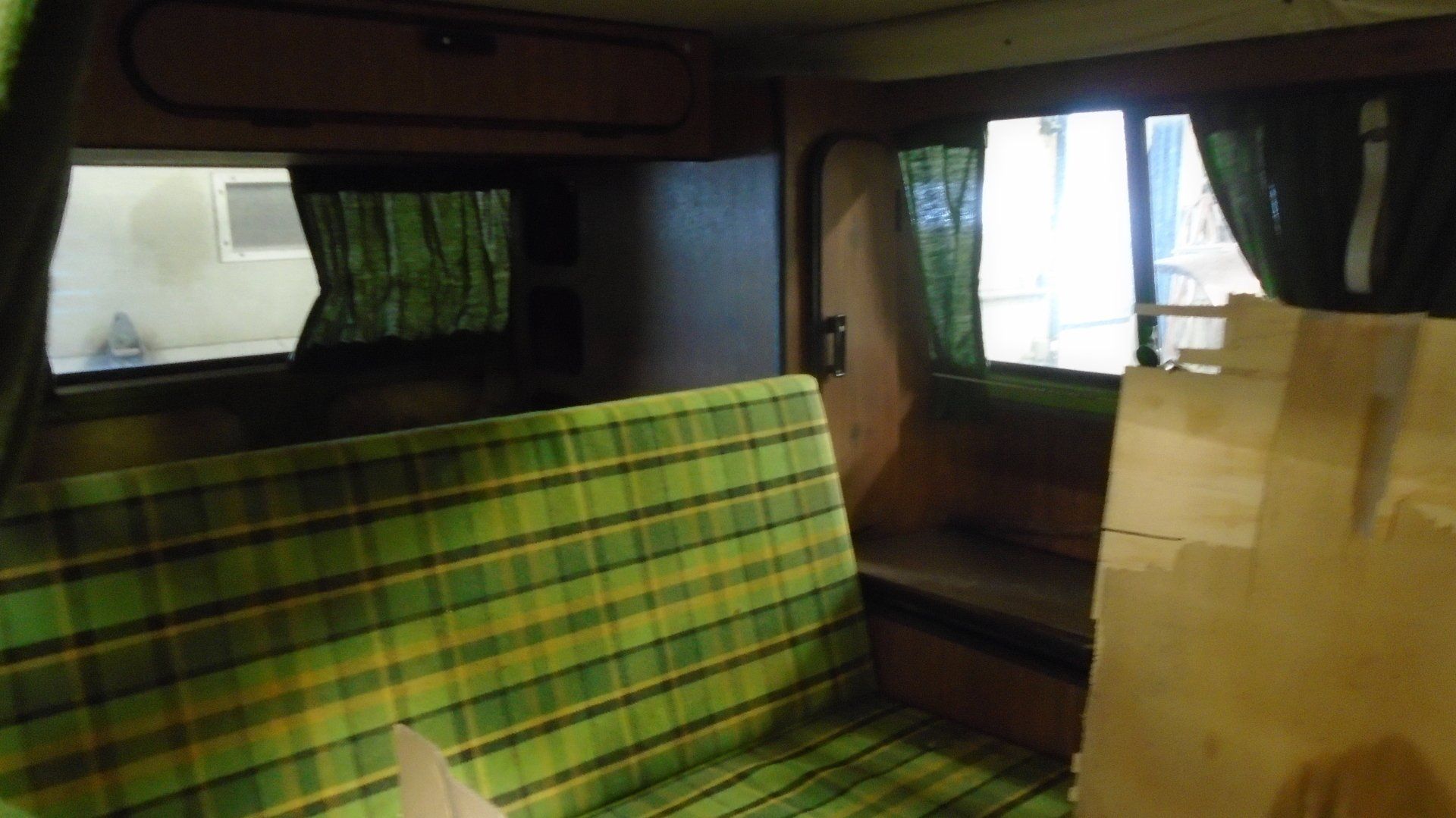 Interior view of a camper van with green plaid seating, small windows, and dark curtains.